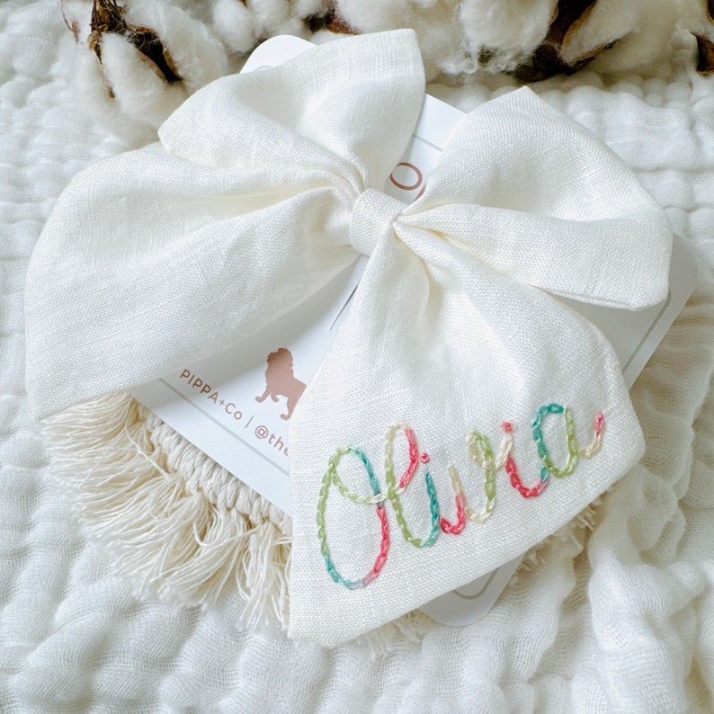 May include: A white linen bow with a white tag that says "PIPPA+CO | @the" and has a colorful embroidered name "Olivia" on it.