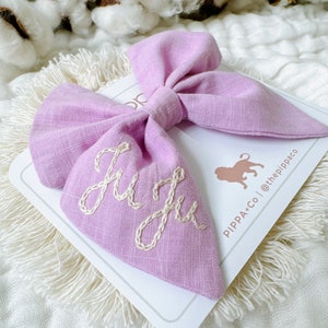May include: A lavender fabric bow with the name "Julia" embroidered in white thread. The bow is on a white card with the text "PIPPA+CO | @thepippaco".