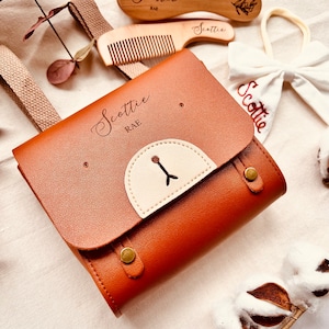 May include: A brown leather baby bag with a white bear face patch and the name "Scottie Rae" engraved on the front. The bag has a strap and a buckle closure.