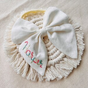May include: A white fabric bow with a yellow headband and a white crocheted circle with fringe. The bow has the name "Harper" embroidered on it in colorful thread.