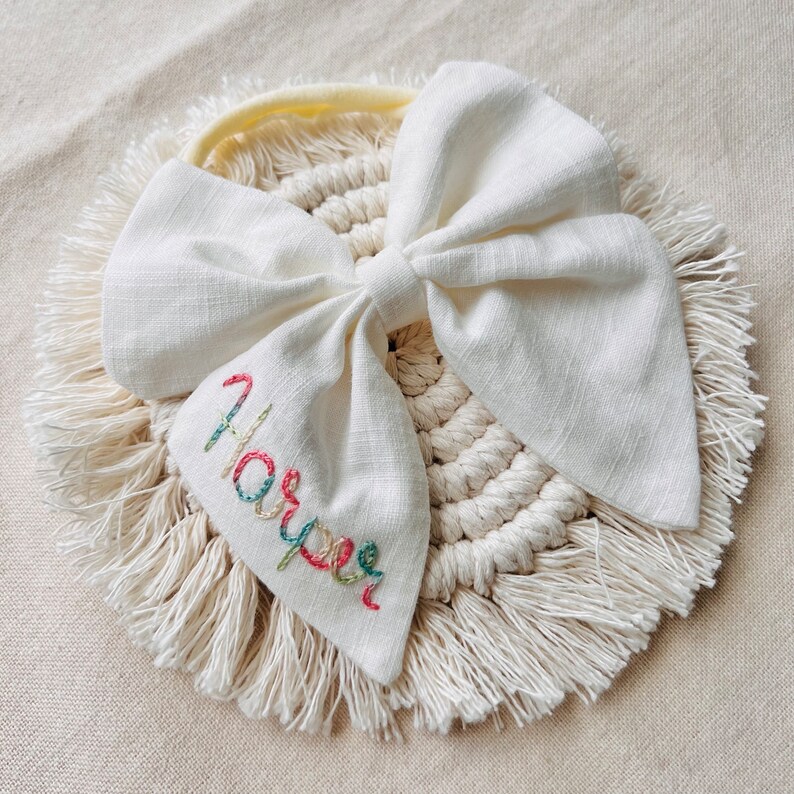 May include: A white fabric bow with a yellow headband and a white crocheted circle with fringe. The bow has the name "Harper" embroidered on it in colorful thread.