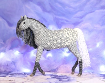 May include: A gray and white crocheted horse toy with a long white tail and mane. The horse is standing on a purple and white background.
