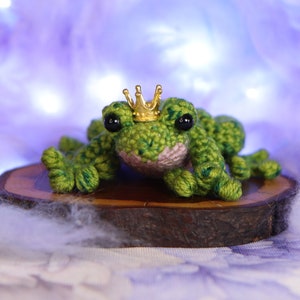 May include: A green crocheted frog wearing a gold crown sits on a wooden slice. The frog has black eyes and a pink belly.