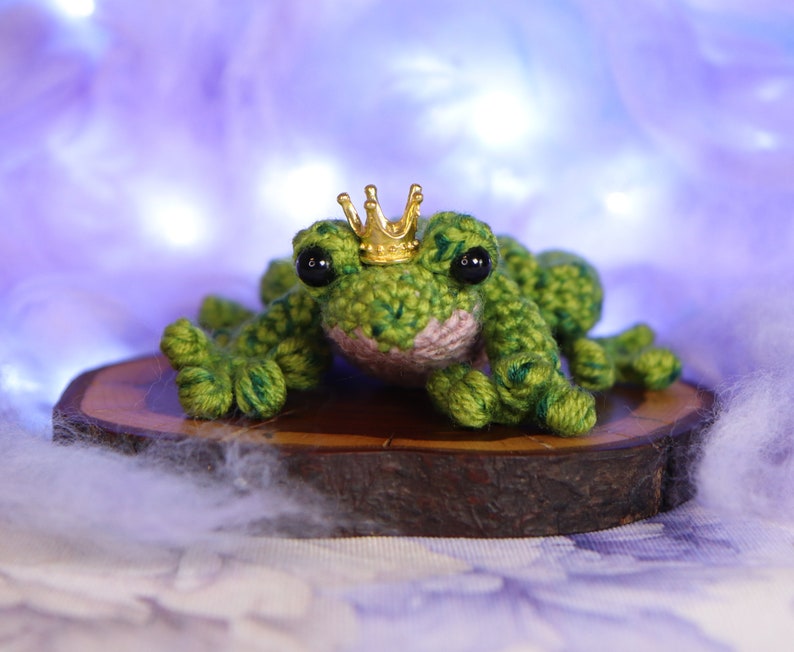 May include: A green crocheted frog wearing a gold crown sits on a wooden slice. The frog has black eyes and a pink belly.