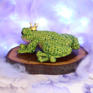 May include: A green crocheted frog wearing a gold crown sits on a wooden slice. The frog is facing away from the camera, with its back legs visible. The frog is sitting on a wooden slice, which is on a white fluffy surface.