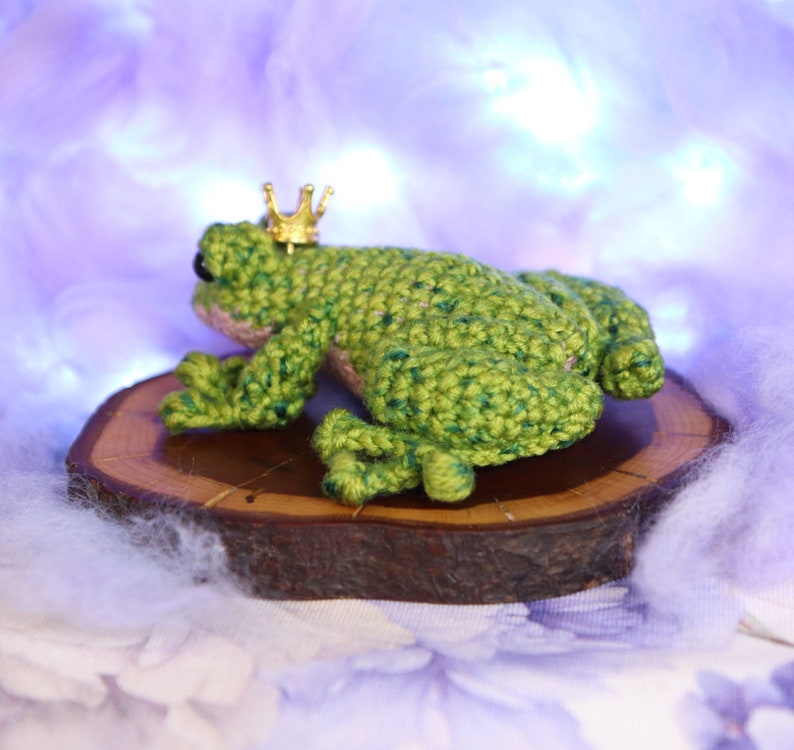 May include: A green crocheted frog wearing a gold crown sits on a wooden slice. The frog is facing away from the camera, with its back legs visible. The frog is sitting on a wooden slice, which is on a white fluffy surface.
