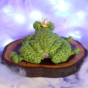 May include: A green crocheted frog wearing a gold crown, sitting on a wooden slice. The frog is lying on its back with its legs in the air.