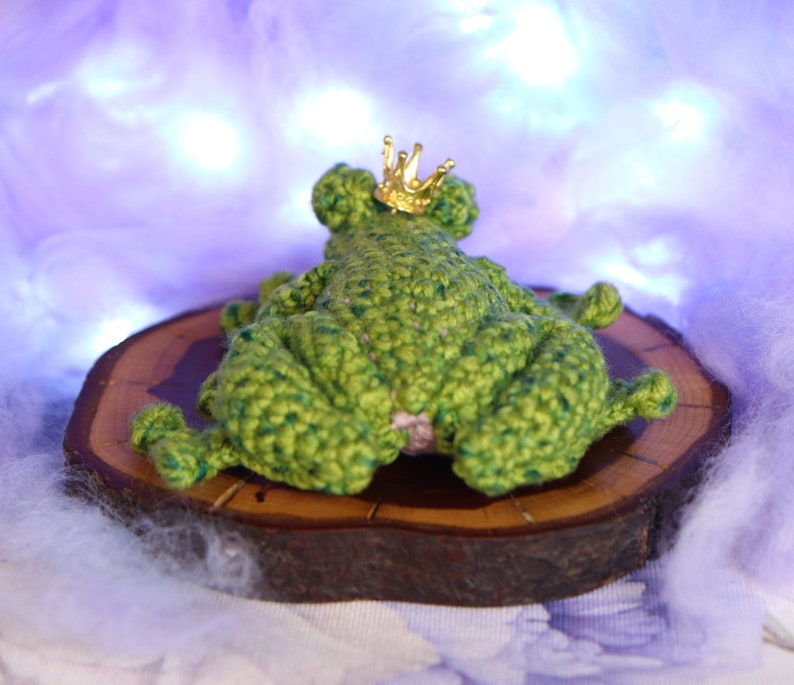 May include: A green crocheted frog wearing a gold crown, sitting on a wooden slice. The frog is lying on its back with its legs in the air.