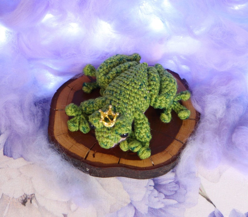 May include: A green crocheted frog wearing a gold crown sits on a wooden slice. The frog is lying on its stomach with its legs outstretched. The background is a soft, purple, fluffy material.