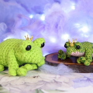 May include: Two crocheted green frog plushies wearing gold crowns. The larger frog is lying down, while the smaller frog is sitting up. Both frogs have black eyes.