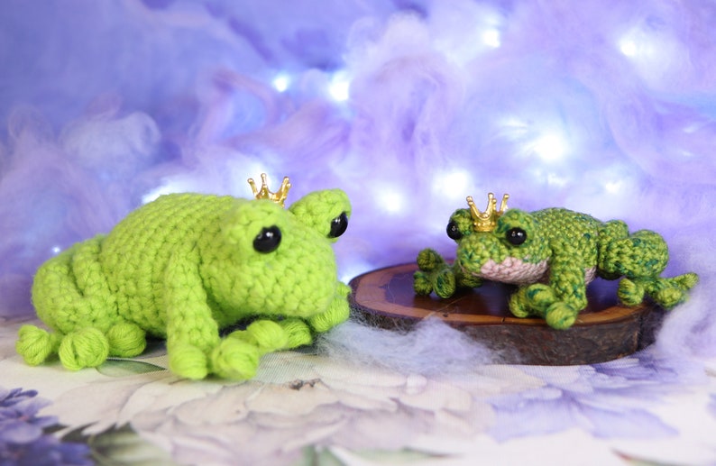 May include: Two crocheted green frog plushies wearing gold crowns. The larger frog is lying down, while the smaller frog is sitting up. Both frogs have black eyes.