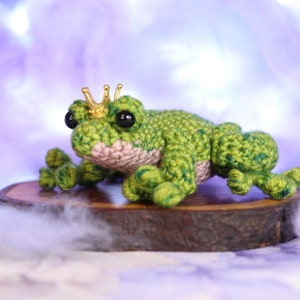 May include: A green crocheted frog wearing a gold crown sits on a wooden log. The frog has black eyes and a pink belly.