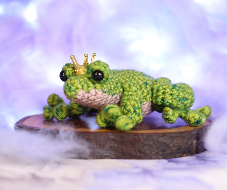 May include: A green crocheted frog wearing a gold crown sits on a wooden log. The frog has black eyes and a pink belly.
