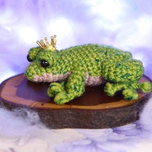 May include: A green crocheted frog wearing a gold crown sits on a wooden slice. The frog has black eyes and a pink belly.