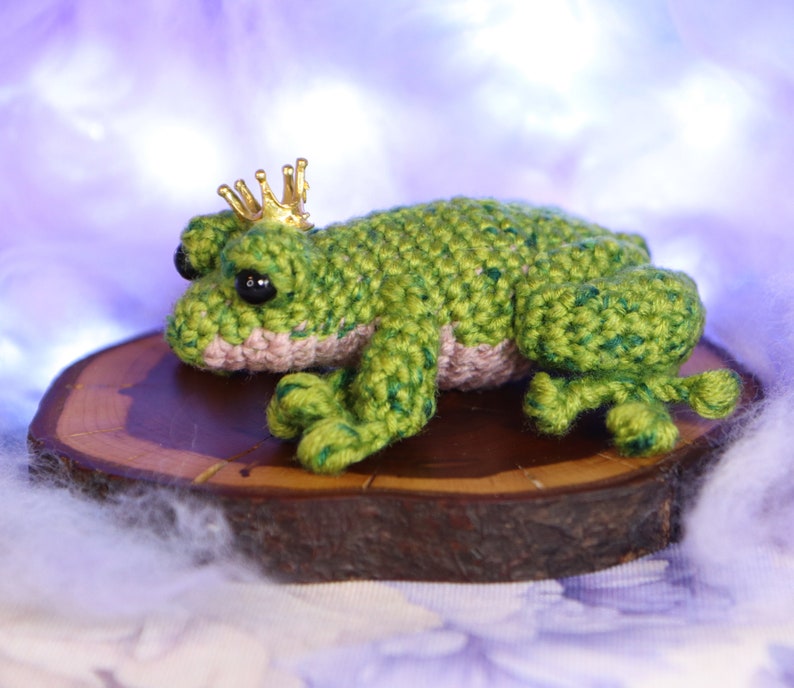 May include: A green crocheted frog wearing a gold crown sits on a wooden slice. The frog has black eyes and a pink belly.
