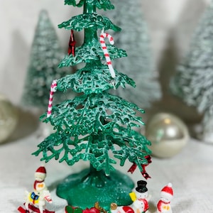 May include: A green Christmas tree with candy cane and star ornaments. Miniature figurines of a snowman, Santa Claus, and a rocking horse are at the base of the tree. A red sleigh is also present, creating a festive holiday scene.