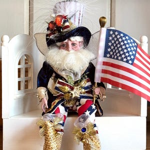 May include: A decorative doll featuring a white beard, wearing a dark blue velvet coat, star-patterned trousers, and a top hat adorned with red, white, and blue accents. The doll holds a miniature American flag and wears gold sequined shoes.