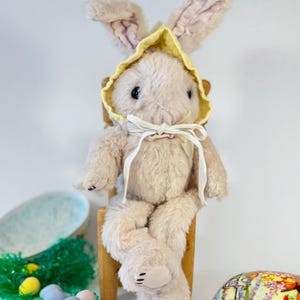 May include: A plush bunny rabbit wearing a yellow bonnet sits on a wooden chair. The bunny is light pink with black eyes and a white ribbon around its neck. There are colorful Easter eggs scattered on the floor in front of the bunny.