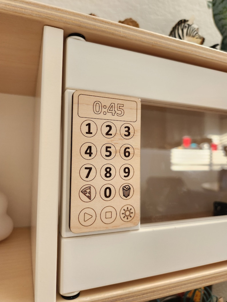 May include: A wooden toy microwave with a digital display showing 0:45. The microwave has a keypad with numbers 1-9 and 0, as well as icons for pizza, popcorn, and a play button.