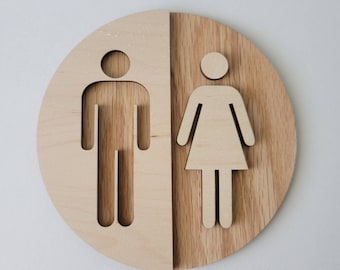 Può includere: Un cartello rotondo in legno con un simbolo maschile e femminile ritagliato. Il cartello è diviso a metà, con il simbolo maschile a sinistra e il simbolo femminile a destra.