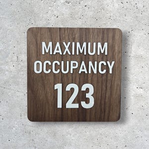 May include: Square wooden sign with the words "MAXIMUM OCCUPANCY 123" in white lettering. The sign has rounded corners and a wood grain pattern. The background is a textured gray wall.