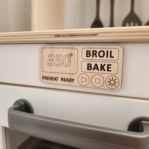 May include: A white toy oven with a wooden control panel. The panel has a digital display showing 350 degrees, buttons for broil and bake, and a preheat ready indicator. The oven has a gray handle and a glass door.