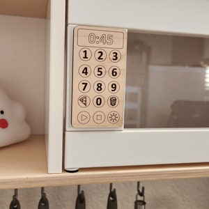 May include: A white toy microwave with a wooden control panel featuring a digital display showing "0:45" and a keypad with numbers 1-9, 0, and icons for pizza, popcorn, play, stop, and a sun.