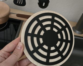 Puede incluir: Un salvamanteles redondo de madera con un diseño geométrico negro. El salvamanteles presenta círculos concéntricos y un centro negro sólido. Utensilios de cocina cuelgan en el fondo. El salvamanteles es sostenido por una mano.