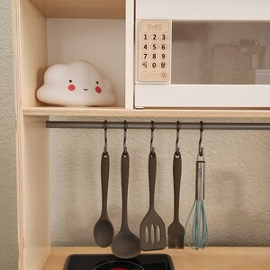 May include: A wooden play kitchen with a black stovetop, a white microwave with a digital display, and a gray metal rack with hooks for hanging utensils. A white cloud nightlight sits on the shelf above the microwave.