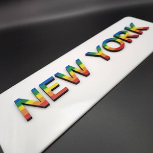 May include: A white rectangular sign with the word "NEW YORK" in rainbow-colored letters.