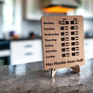 May include: A wooden dog feeding tracker with a stand. The tracker has a list of days of the week with checkboxes for AM and PM feeding times. The text "Has Hunter been fed?" is printed at the bottom.
