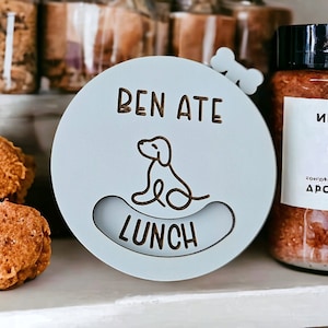 May include: A light blue round wooden sign with the text "Ben Ate Lunch" and a cartoon dog sitting next to a bone. The sign is on a white shelf with other items.
