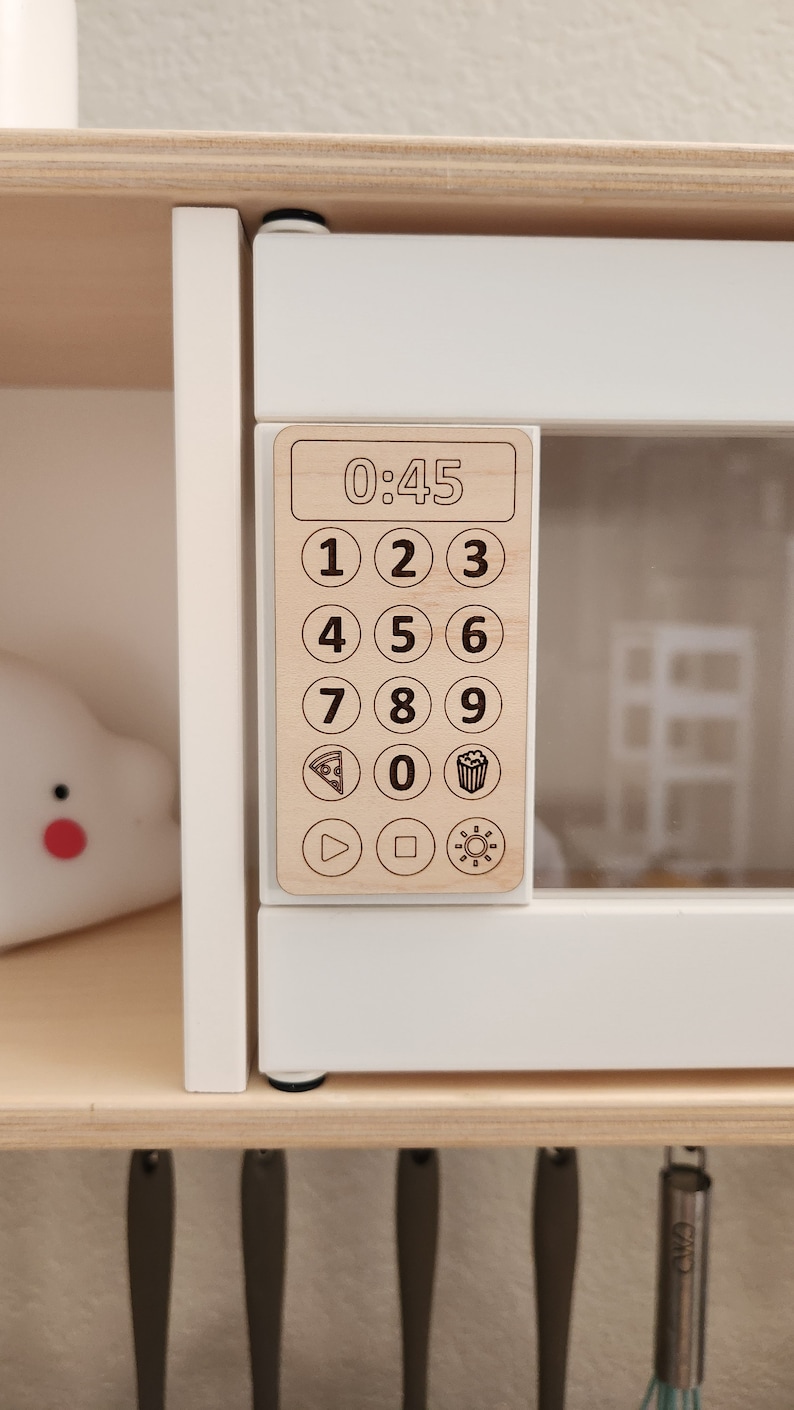 May include: A white toy microwave with a wooden control panel featuring a digital timer display showing 0:45, number buttons, and icons for pizza, popcorn, and a play button.