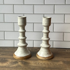 Könnte beinhalten: Zwei weiße Kerzenhalter aus Keramik mit gesprenkeltem Finish und braunem Sockel. Die Kerzenhalter haben ein einzigartiges, strukturiertes Design.