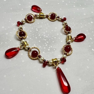 May include: A gold bracelet with red beads and teardrop charms. The bracelet also features small white pearls and gold accents. The red beads are set in gold circular frames. The bracelet is approximately 18 cm in length.