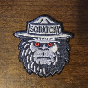 May include: A gray and white embroidered patch of a Bigfoot wearing a ranger hat with the word "Squatchy" on the hat.
