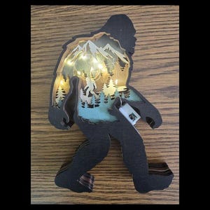 May include: A wooden wall decor featuring a silhouette of Bigfoot with a layered design of a mountain scene and forest inside. The silhouette is black and the mountain scene is in shades of brown, blue, and green. There are small white lights inside the mountain scene.
