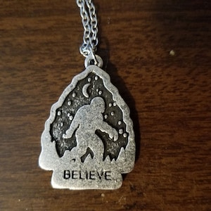 May include: A silver necklace with a pendant featuring a silhouette of Bigfoot walking in a forest with a crescent moon and stars. The pendant has the word "BELIEVE" inscribed on the bottom.