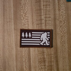 May include: A rectangular patch with a dark brown border and a dark brown background. The design features white horizontal stripes, three white pine trees in the upper left corner, and a white silhouette of a Bigfoot figure on the right side.