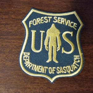 May include: A green and gold embroidered patch with the text "Forest Service" and "U.S. Department of Sasquatch" around a silhouette of a sasquatch.