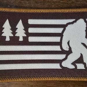 May include: A rectangular patch featuring a brown background with white stripes and three white pine trees. A white silhouette of Bigfoot is on the right side. The patch has a gold border.