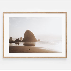May include: A framed photograph of a rocky coastline with a large rock formation in the distance. The beach is sandy and the water is calm and reflective. Two people are walking along the beach in the foreground.
