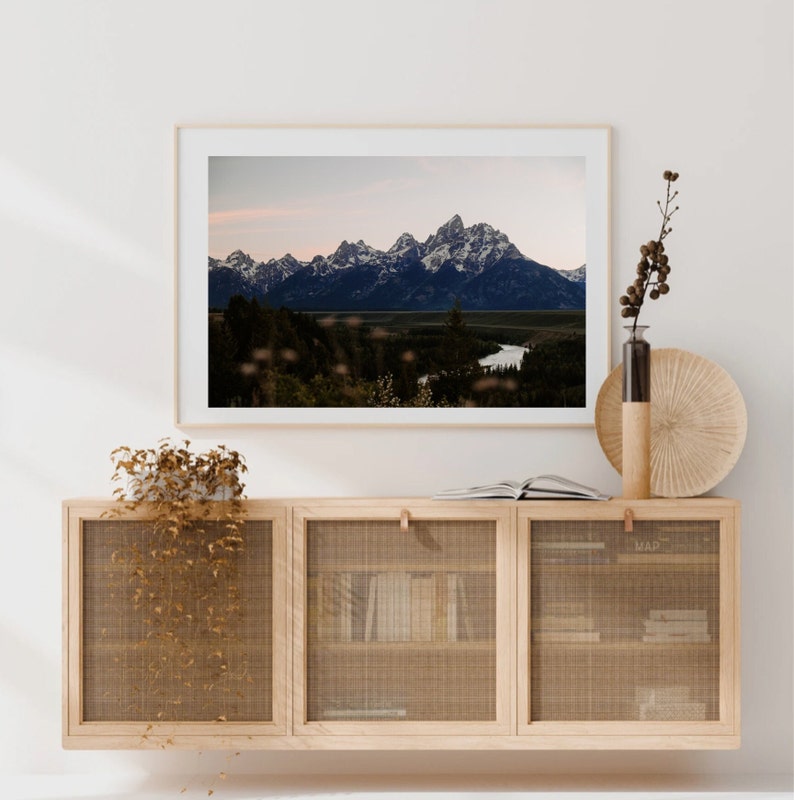 May include: A framed photograph of a mountain range with a river winding through the valley. The mountains are snow-capped and the sky is a soft blue. The photo is hanging on a white wall above a wooden cabinet with a woven mesh front.