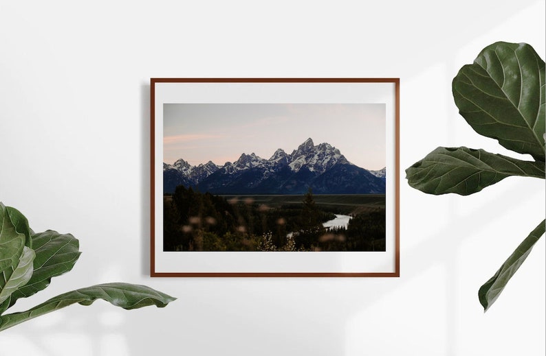 May include: A framed photograph of a mountain range with snow-capped peaks, a river winding through a valley, and a forest in the foreground. The sky is a soft blue with a hint of pink.