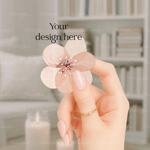 May include: A hand with a gold ring holding a watercolor flower. The flower is peach and white with a detailed center. The text "Your design here" is above the flower.