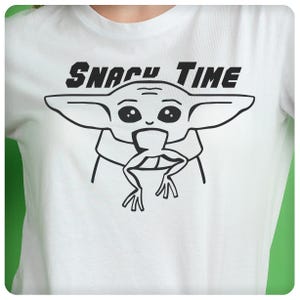 May include: White t-shirt with a black and white illustration of a character from the Star Wars franchise, known as "Baby Yoda", eating a frog. The text "Snacky Time" is printed above the illustration.