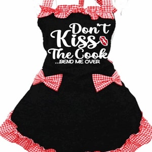 Puede incluir: Delantal negro con ribete a cuadros rojos y blancos. El delantal tiene un gr&aacute;fico blanco que dice "Don't Kiss The Cook...Bend Me Over" con un gr&aacute;fico de labios rojos.