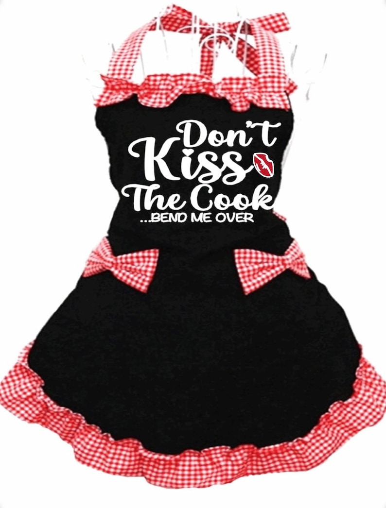 Puede incluir: Delantal negro con ribete a cuadros rojos y blancos. El delantal tiene un gr&aacute;fico blanco que dice "Don't Kiss The Cook...Bend Me Over" con un gr&aacute;fico de labios rojos.