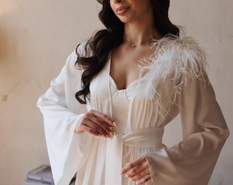 May include: A white satin robe with wide sleeves and a matching belt. The robe features a feather accent on the shoulder. The model is wearing a white slip dress underneath. The background is a neutral colour.