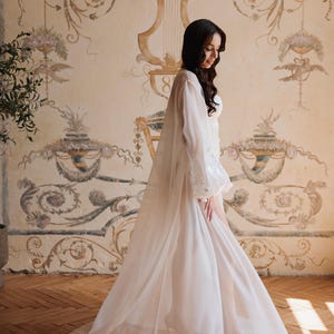 May include: A long, white sheer robe with sleeves and lace detailing. The robe is worn over a white garment. The background features a vintage-style wall mural with floral and scroll designs, and a wooden floor.